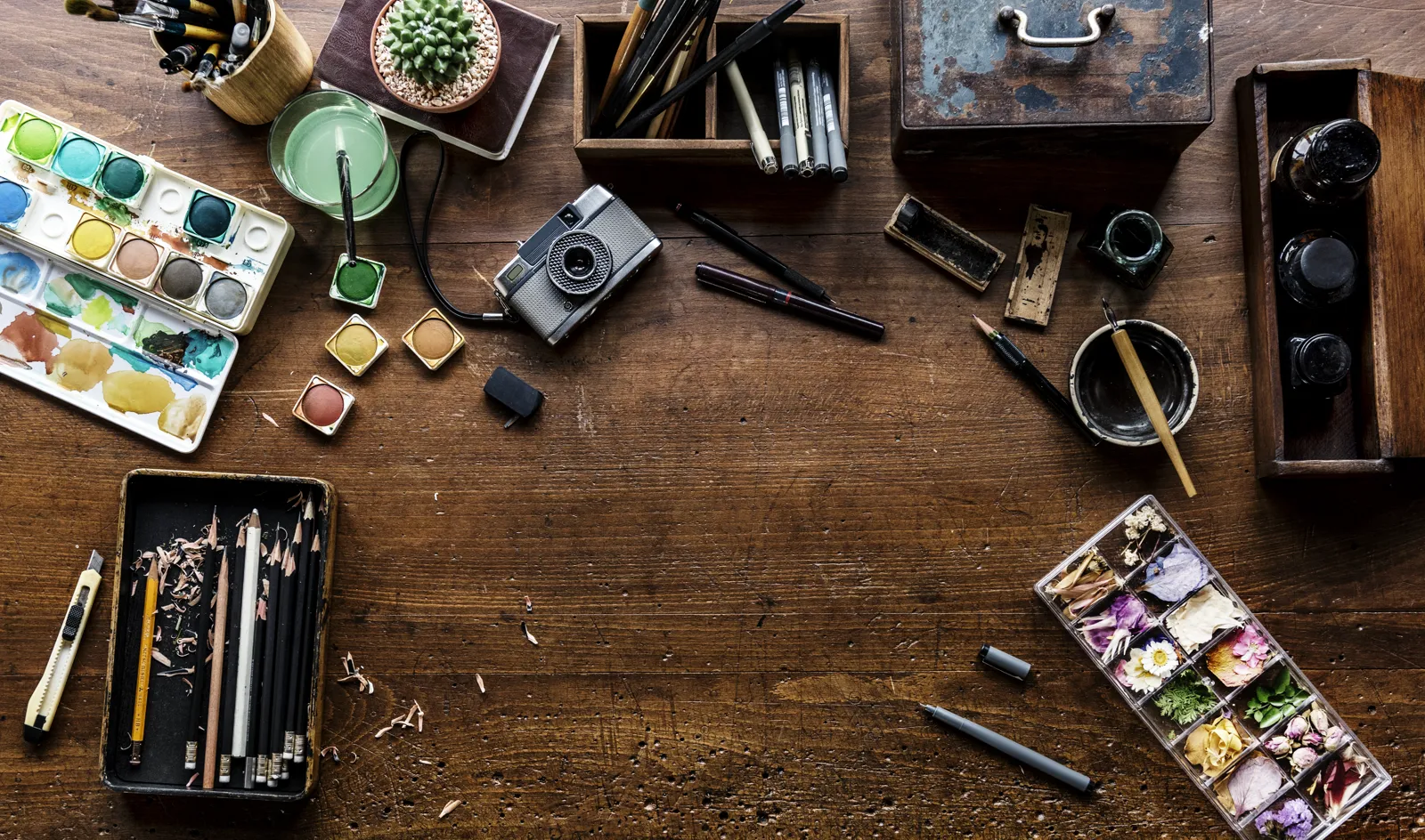 Overhead flat-lay of artistic supplies: paint tubes, brushes, sketchbook, palette, scissors, a small plant