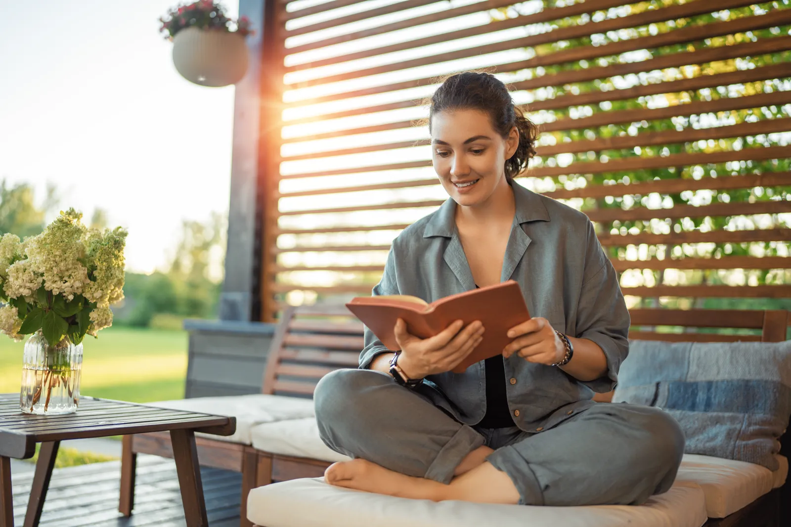A woman reading a book outdoors on a sunny suburban patio, blanket on her lap, slow afternoon energy