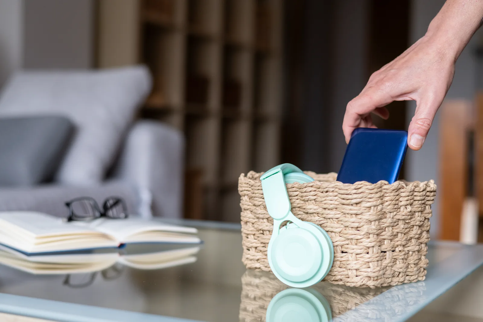 A hand placing a smartphone into a woven basket next to a pair of headphones, soft natural light