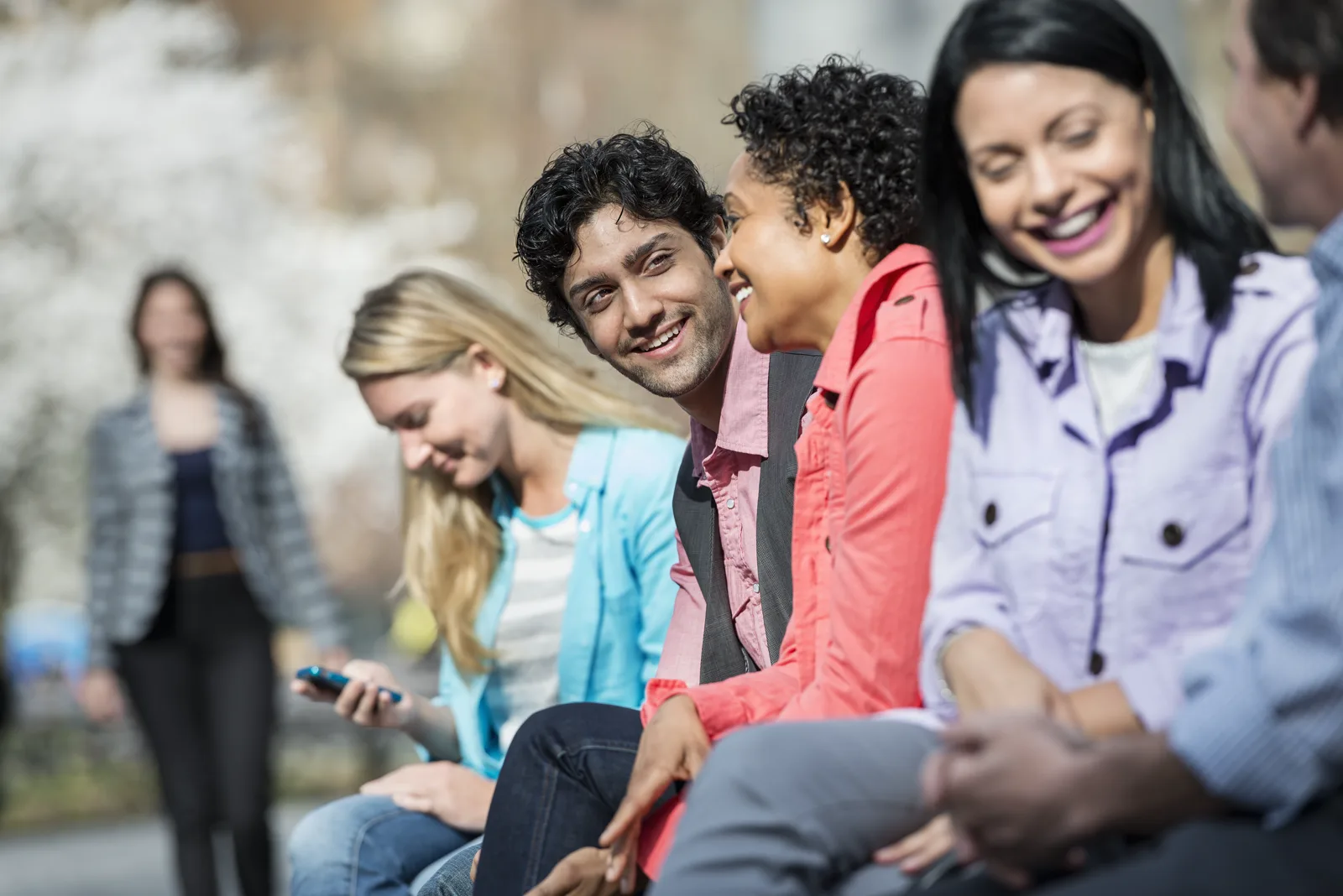Five people sitting on a bench in a park, four of them present with each other while one looks at a smartphone