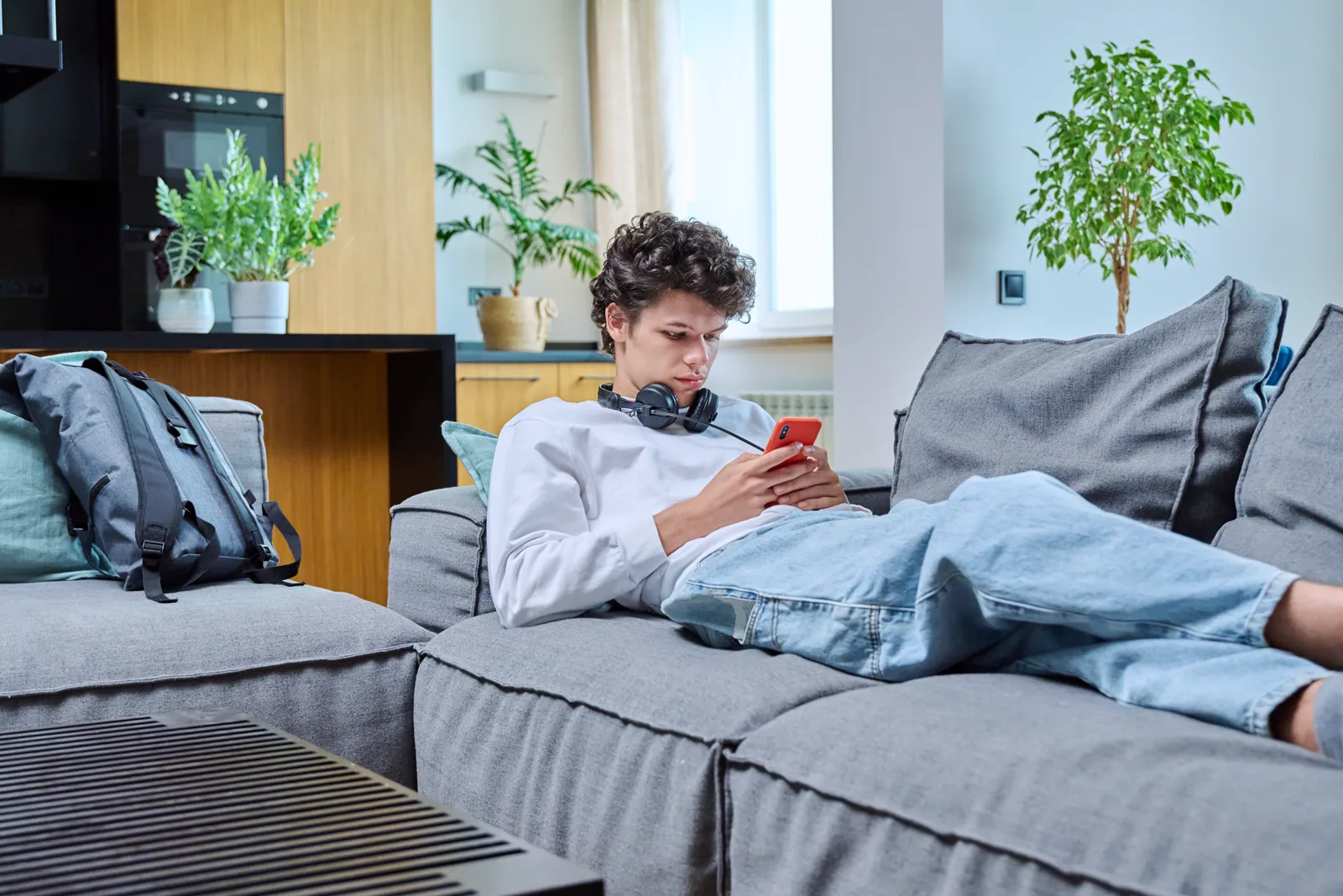 A young person relaxing on a sofa at home with headphones on, using a smartphone, soft natural light