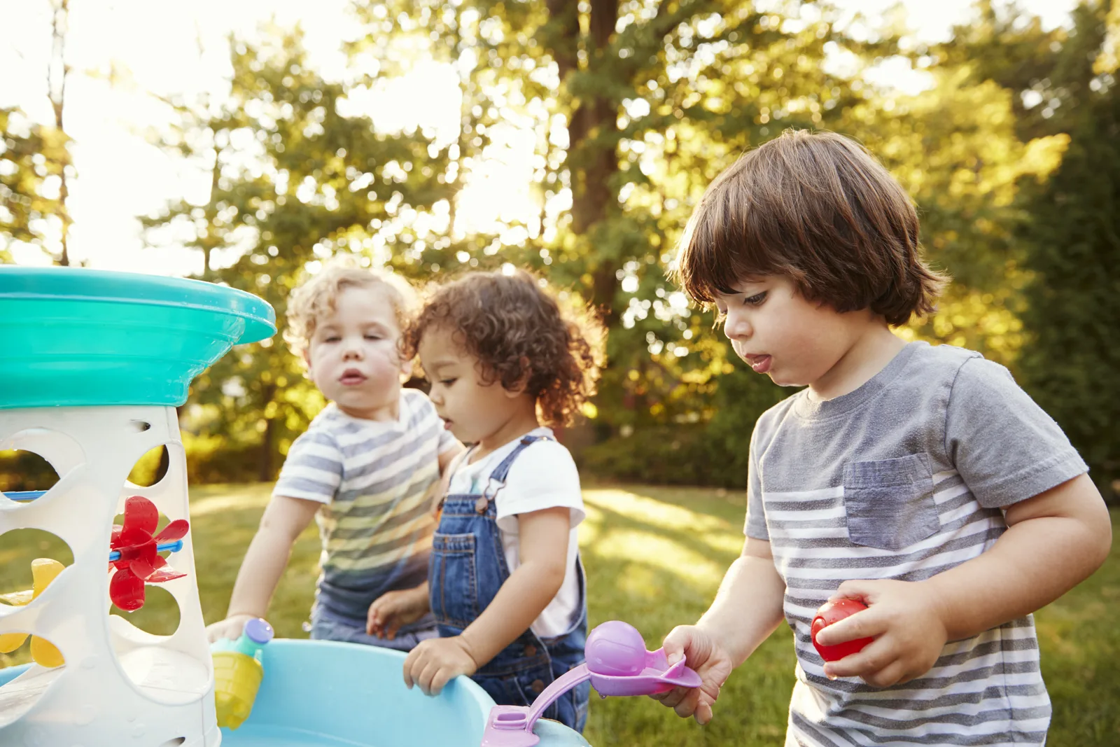 A group of young children gathered around an outdoor water play table, splashing and exploring with their hands