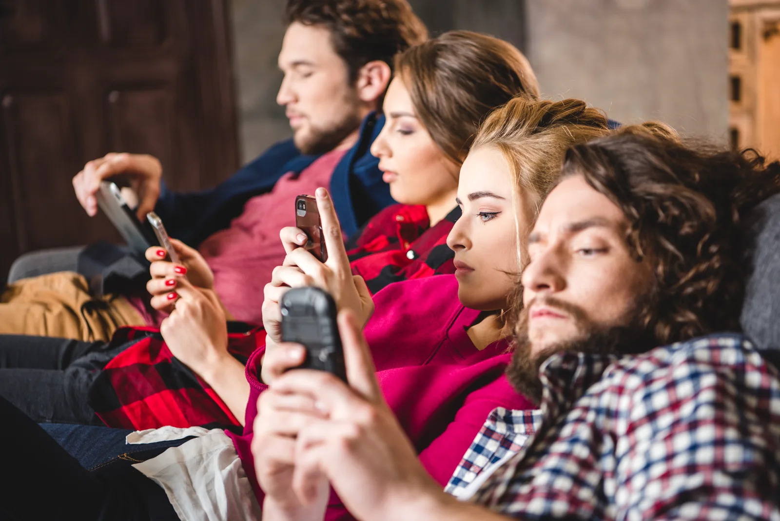 A group of friends sitting close together but each looking down at their own smartphones, side view in soft light