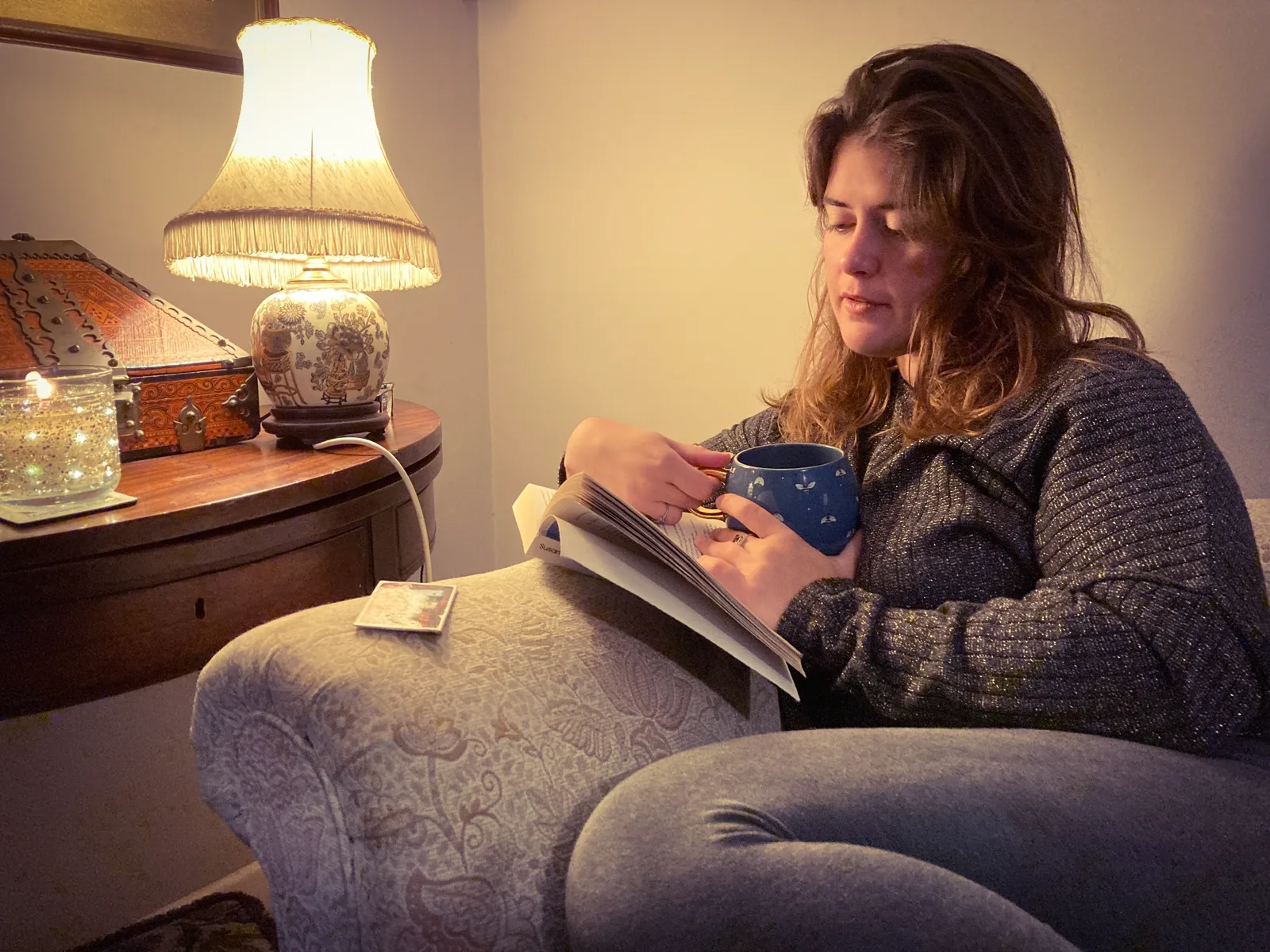 A woman relaxing indoors in warm afternoon light, reading a book with a drink on the table beside her, no phone in sight
