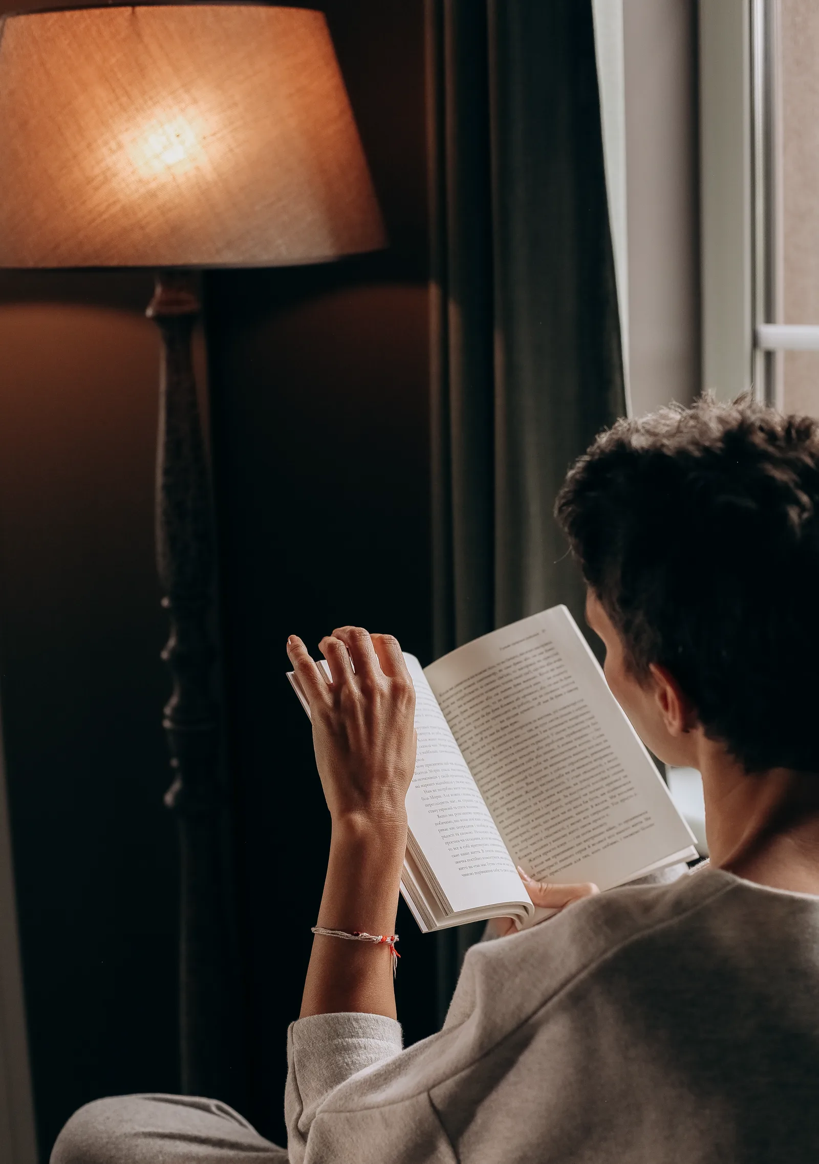 A person settled into a comfortable chair reading a paper book by a single warm lamp in the evening