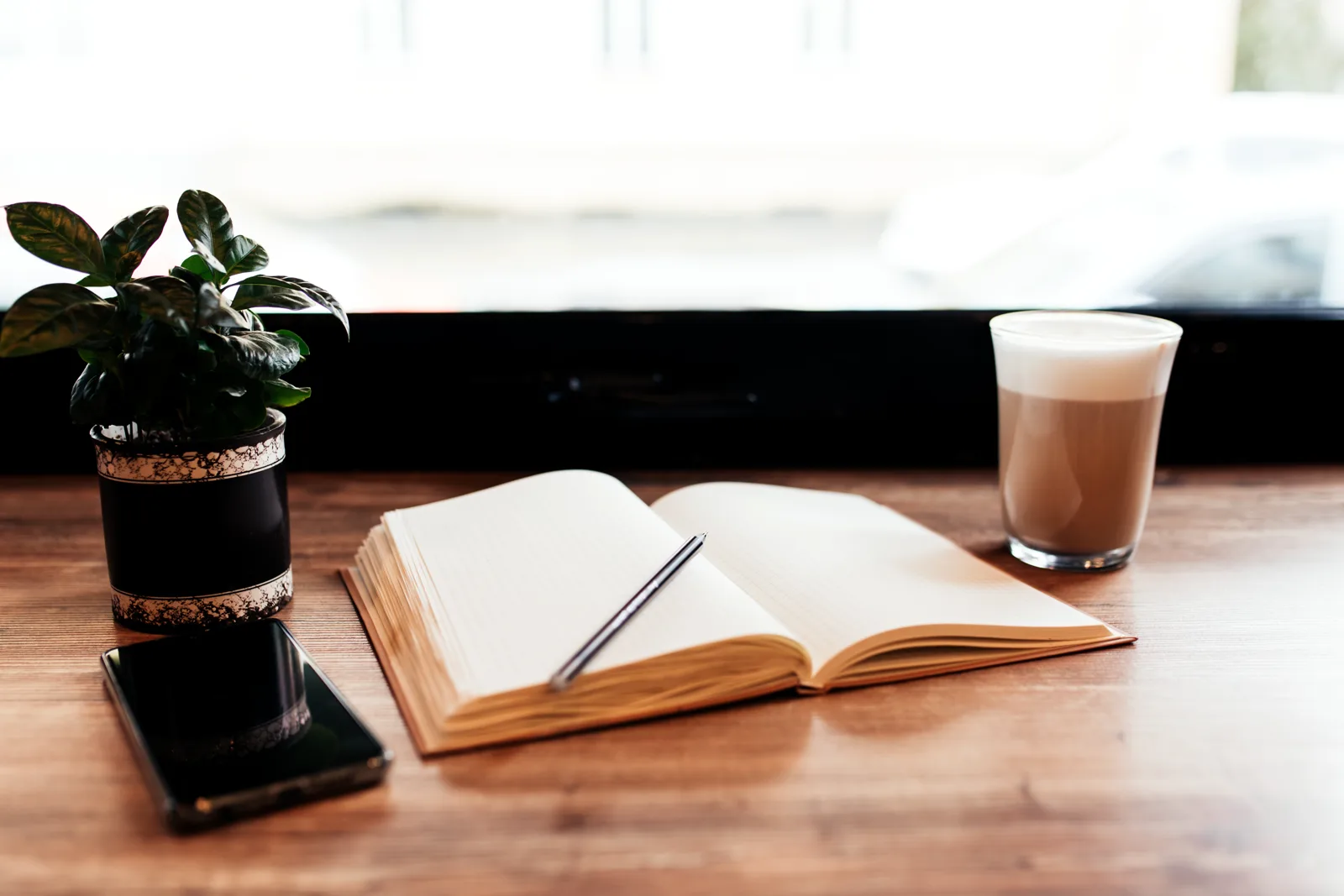 A cup of coffee next to a notebook and a phone on a cafe table, warm morning light