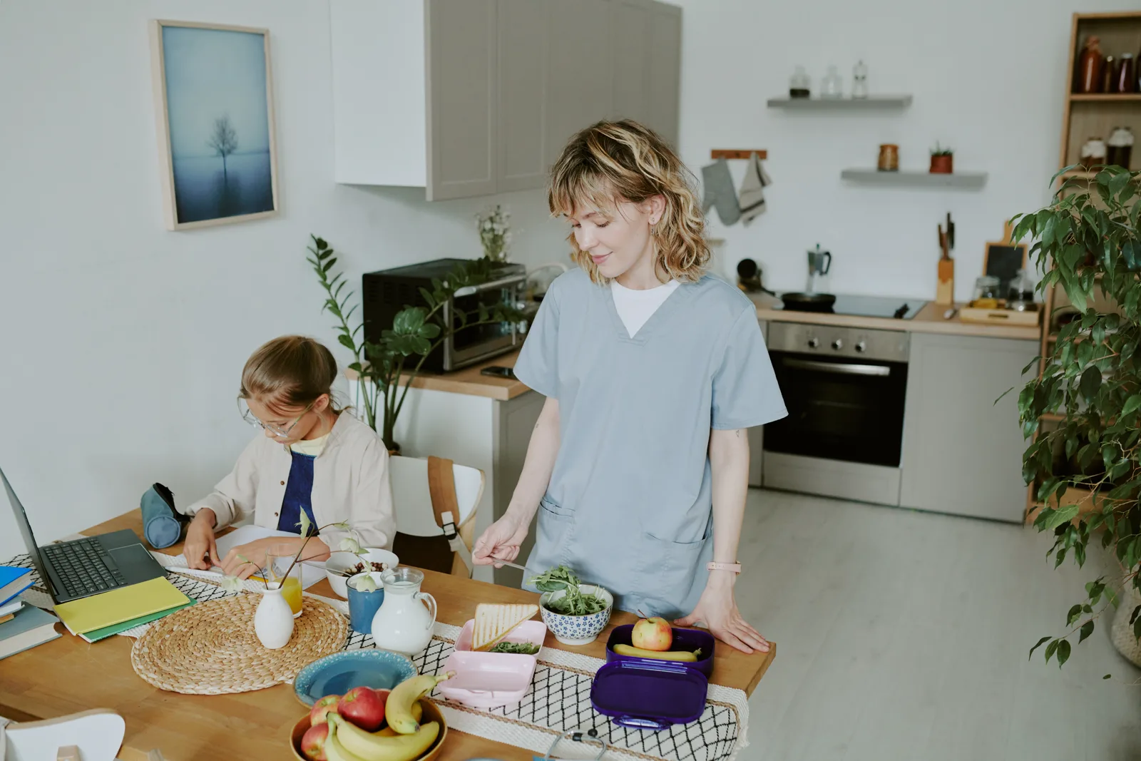 A parent at a kitchen counter managing several things at once in soft daylight