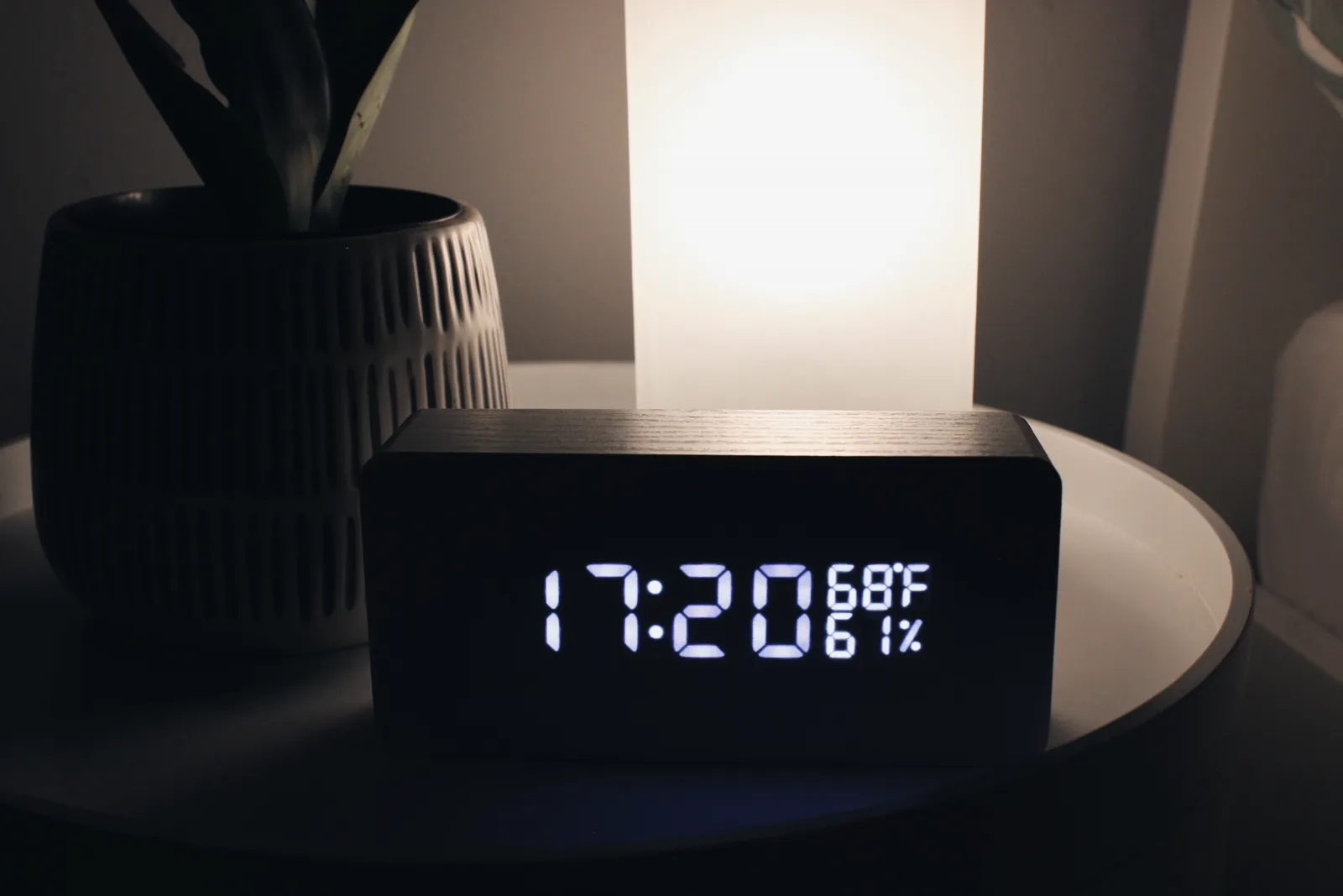 A simple alarm clock on a wooden nightstand beside a book and a small lamp