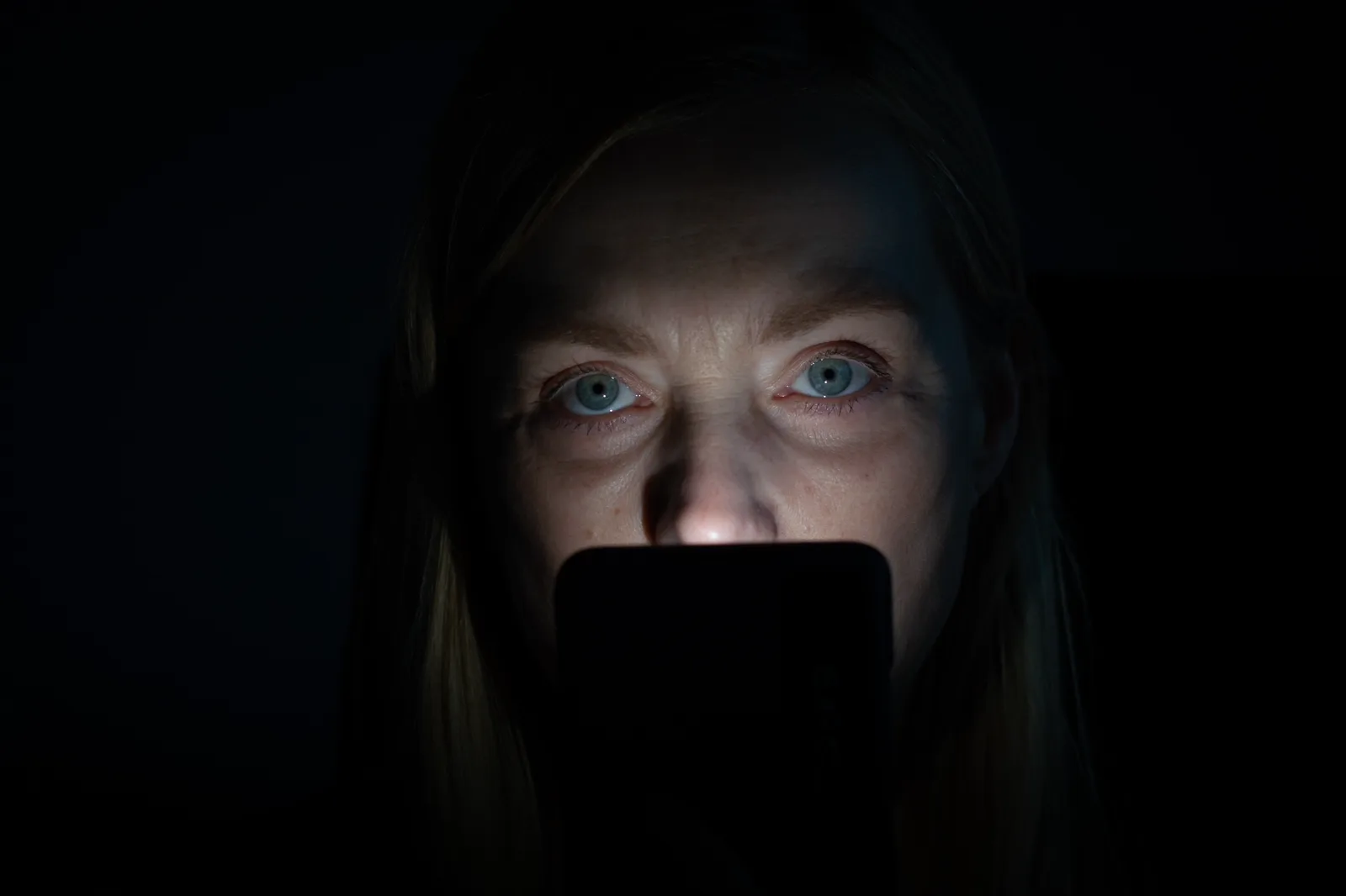 A person's face lit by a phone screen in the dark, absorbed in an endless feed