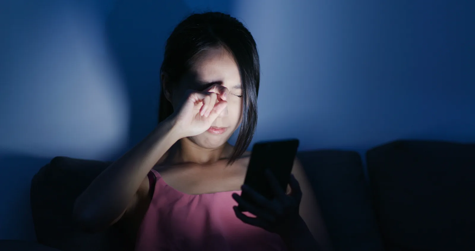 A tired woman rubbing her eyes while looking at her smartphone, low light