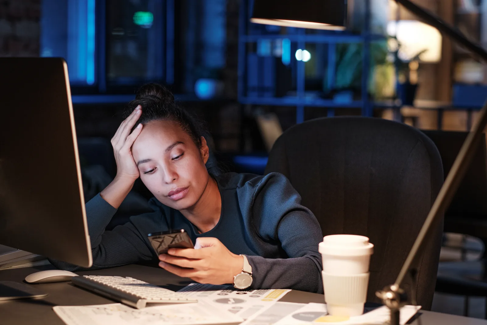 An exhausted young woman sitting at a desk at night, leaning her head on her hand while checking her phone